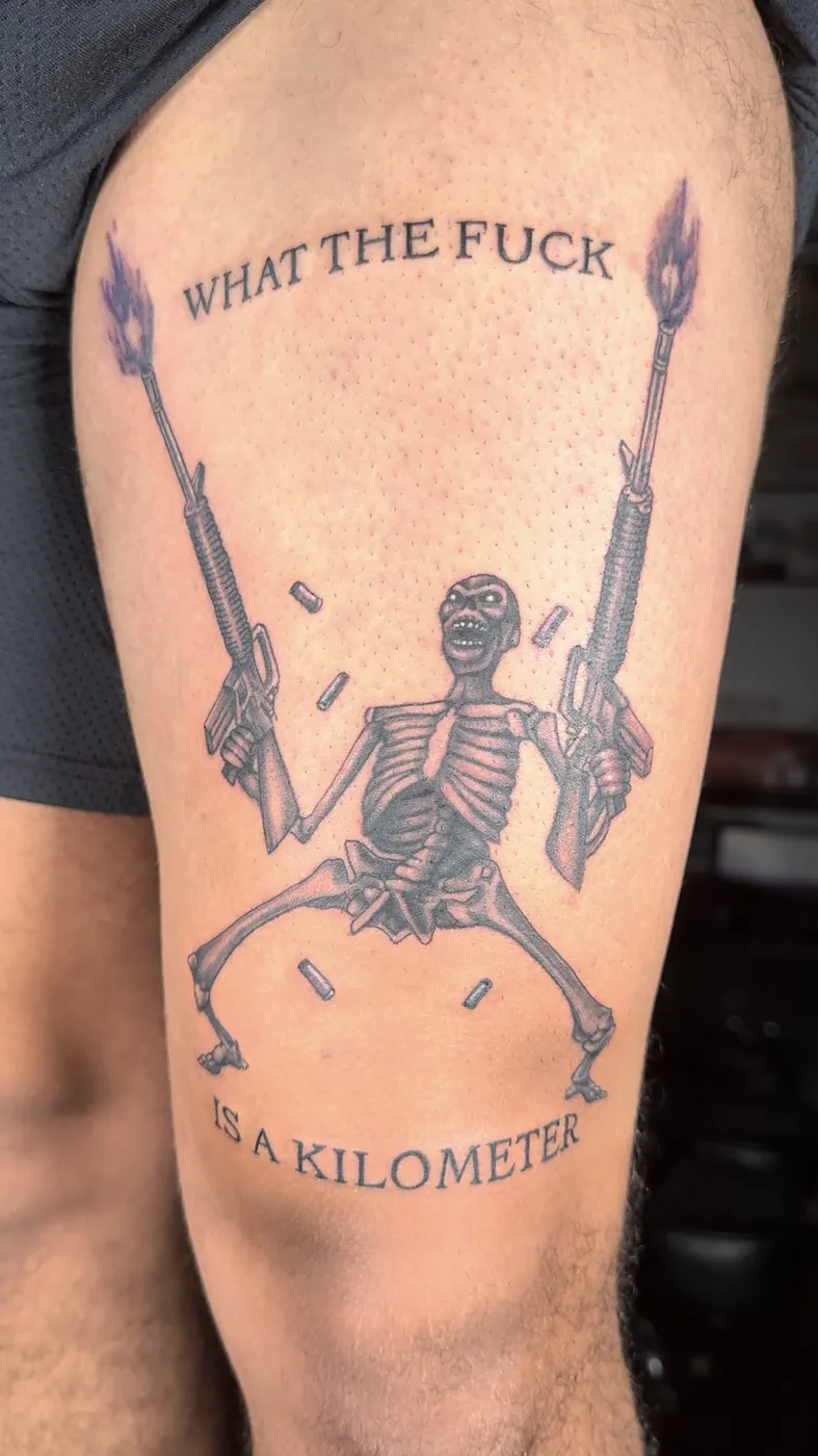 Squelette humoristique tenant deux fusils, tatoué sur la cuisse, couleur.